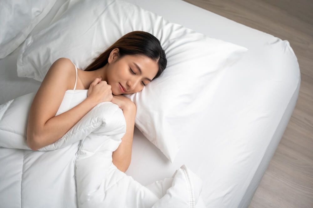 cách làm mặt thon gọn cô gái đang nhắm mắt và đắp chăn nằm trên tấm đệm màu trắng