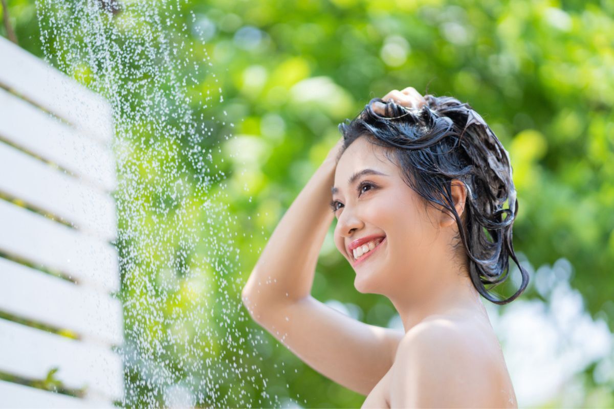gội đầu nhiều có tốt không người phụ nữ châu Á tắm ngoài trời đang dùng tay chải tóc và mỉm cười.