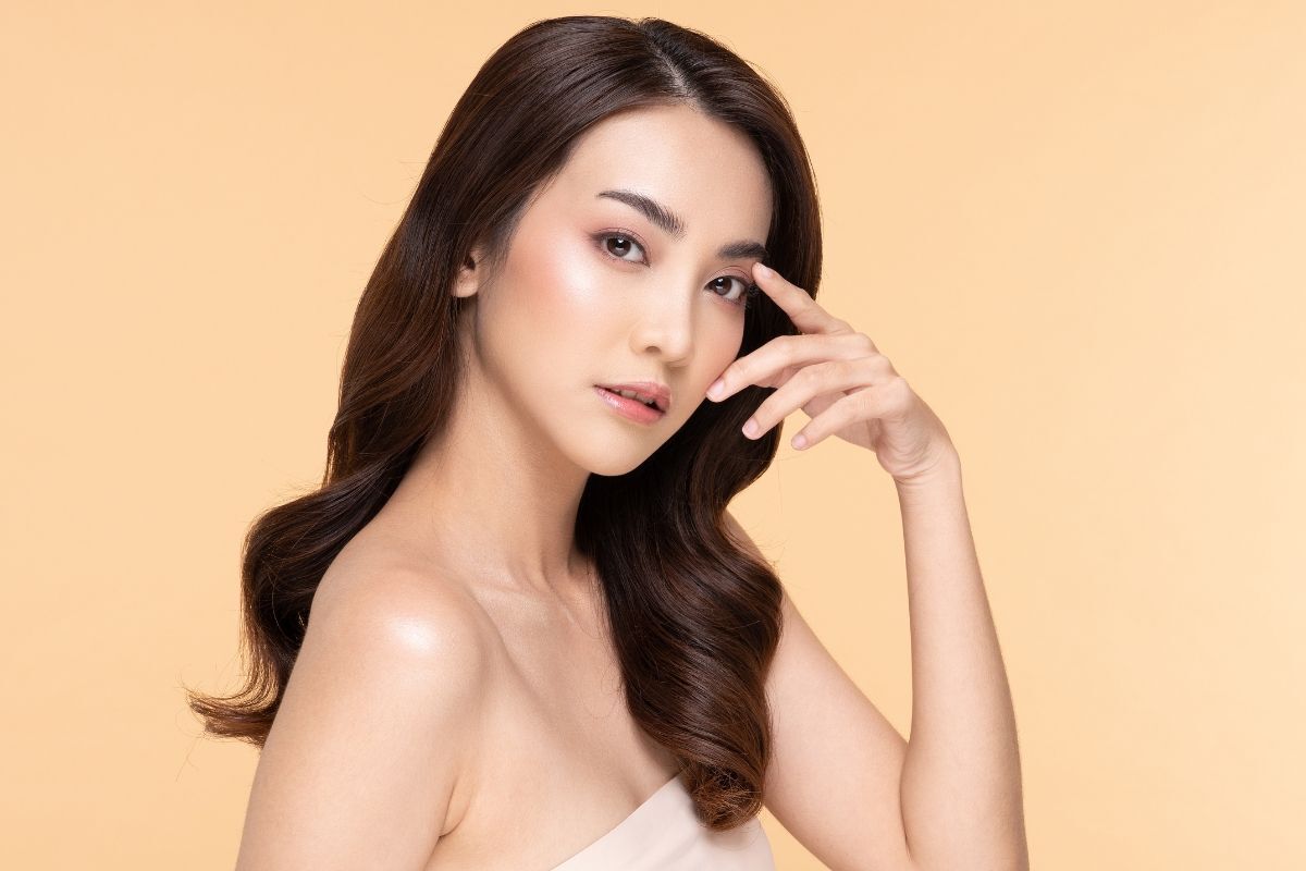 dáng lông mày đẹp cô gái tóc xoăn nhẹ trang điểm tông cam lấy hai ngón tay để lên mặt ngay vùng mắt trên nền cam nhạt 