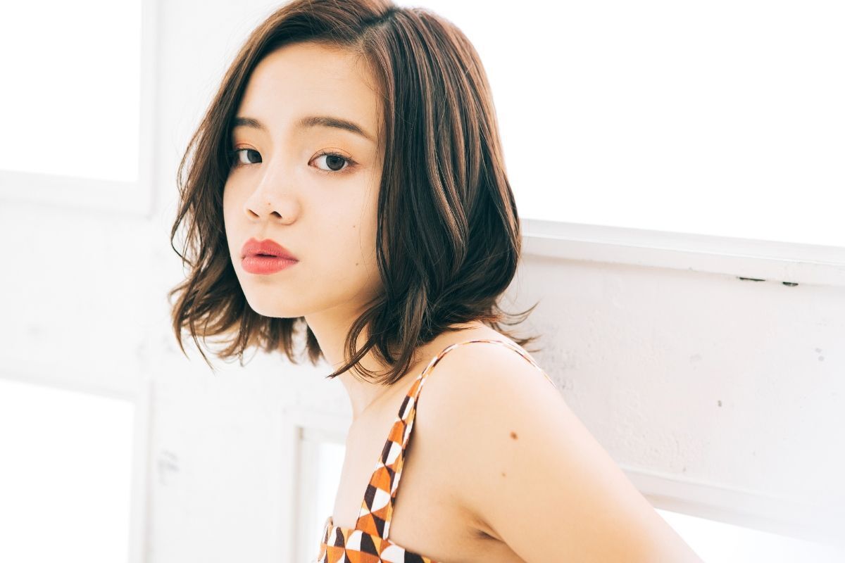Tóc ngắn ngang vai uốn gợn sóng nhẹ góc nghiêng của cô gái mặc váy đang đứng trước ô cửa sổ có ánh nắng chiếu vào ảnh chụp trong phòng