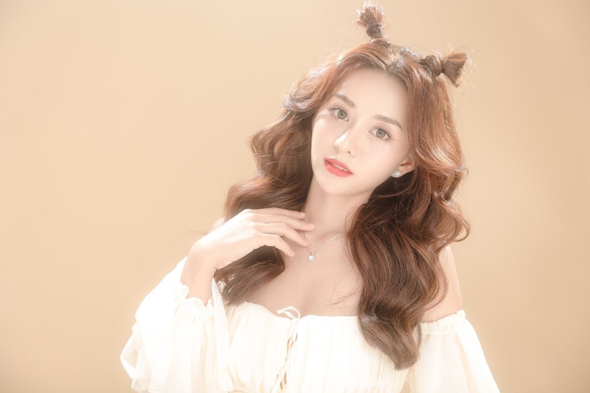 Tóc xoăn sóng nước cho mặt dài cô gái búi tóc hai bên mặc áo trắng trễ vai chụp hình trên phông nền màu nâu be