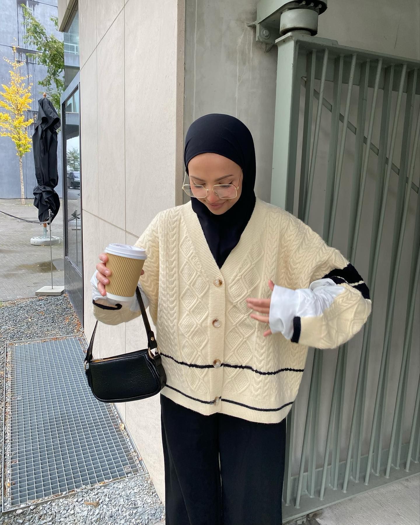 Wanita Kaukasia mengenakan hijab warna hitam dan atasan knit warna cream sambil memegang segelas kopi.