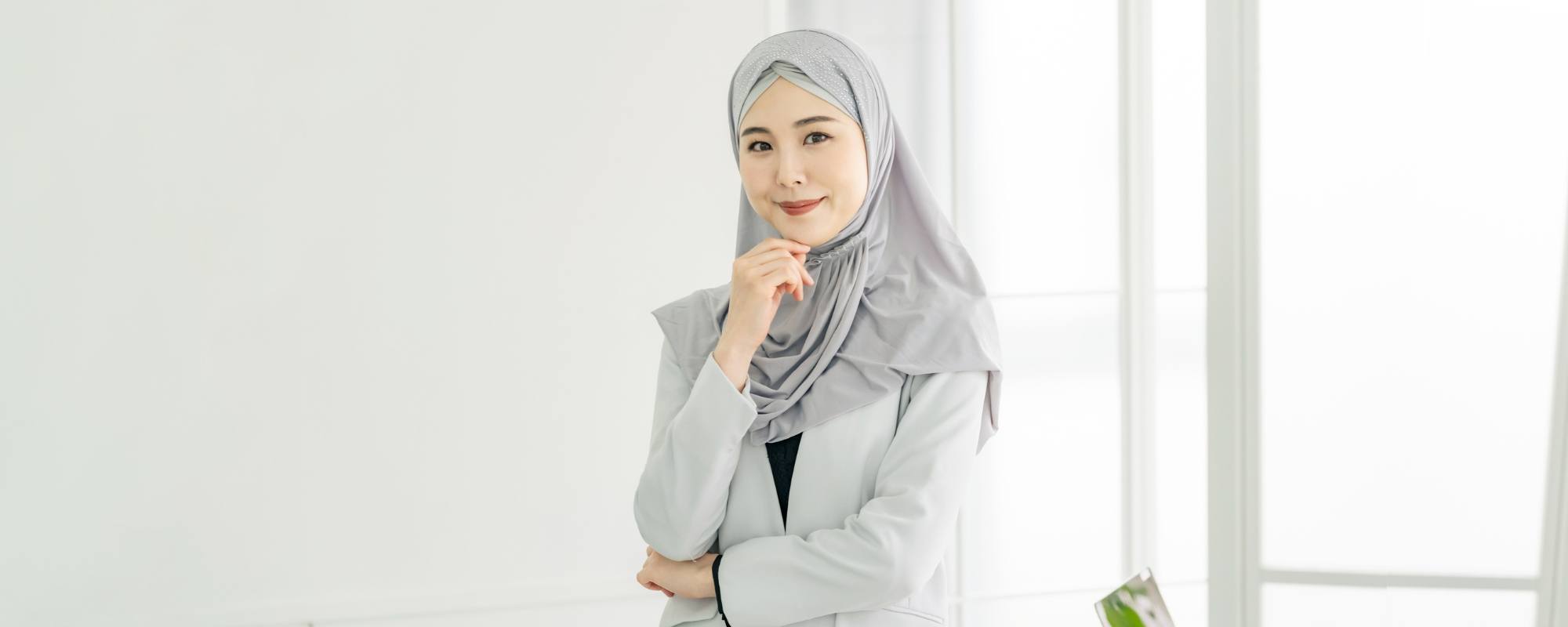 Wanita Asia sedang berpangku tangan mengenakan hijab warna abu-abu dan blazer warna putih.
