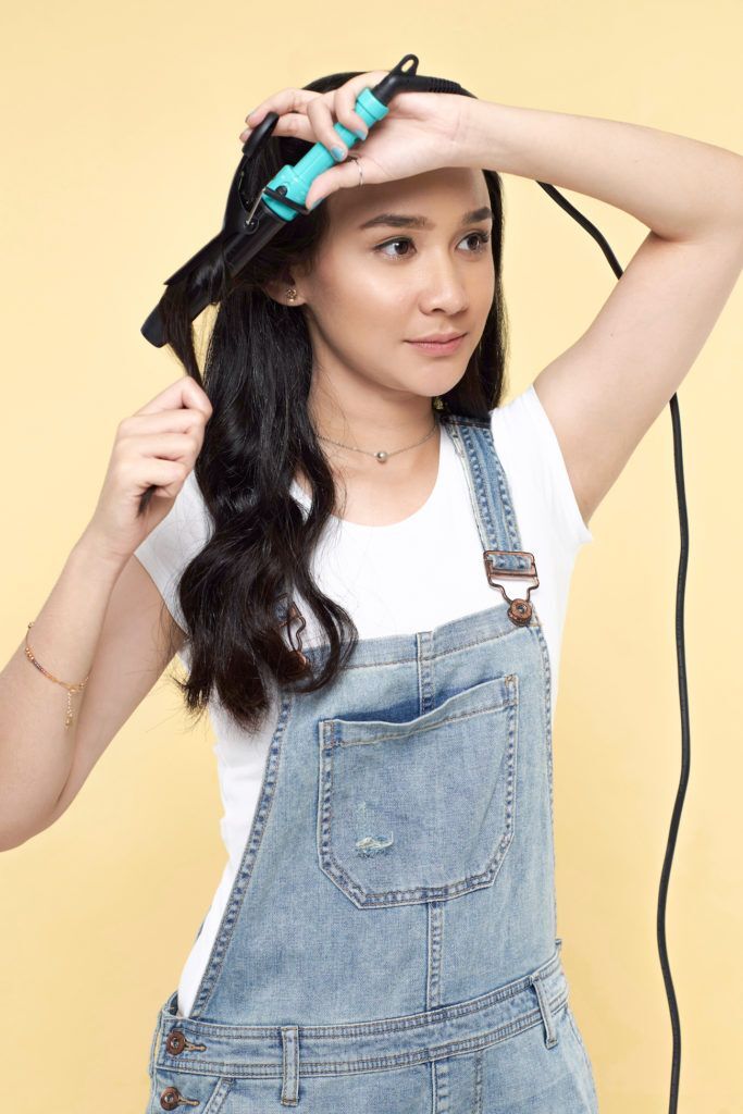 Wanita asia dengan rambut hitam panjang gaya wavy sedang mengeriting rambut dengan curling iron