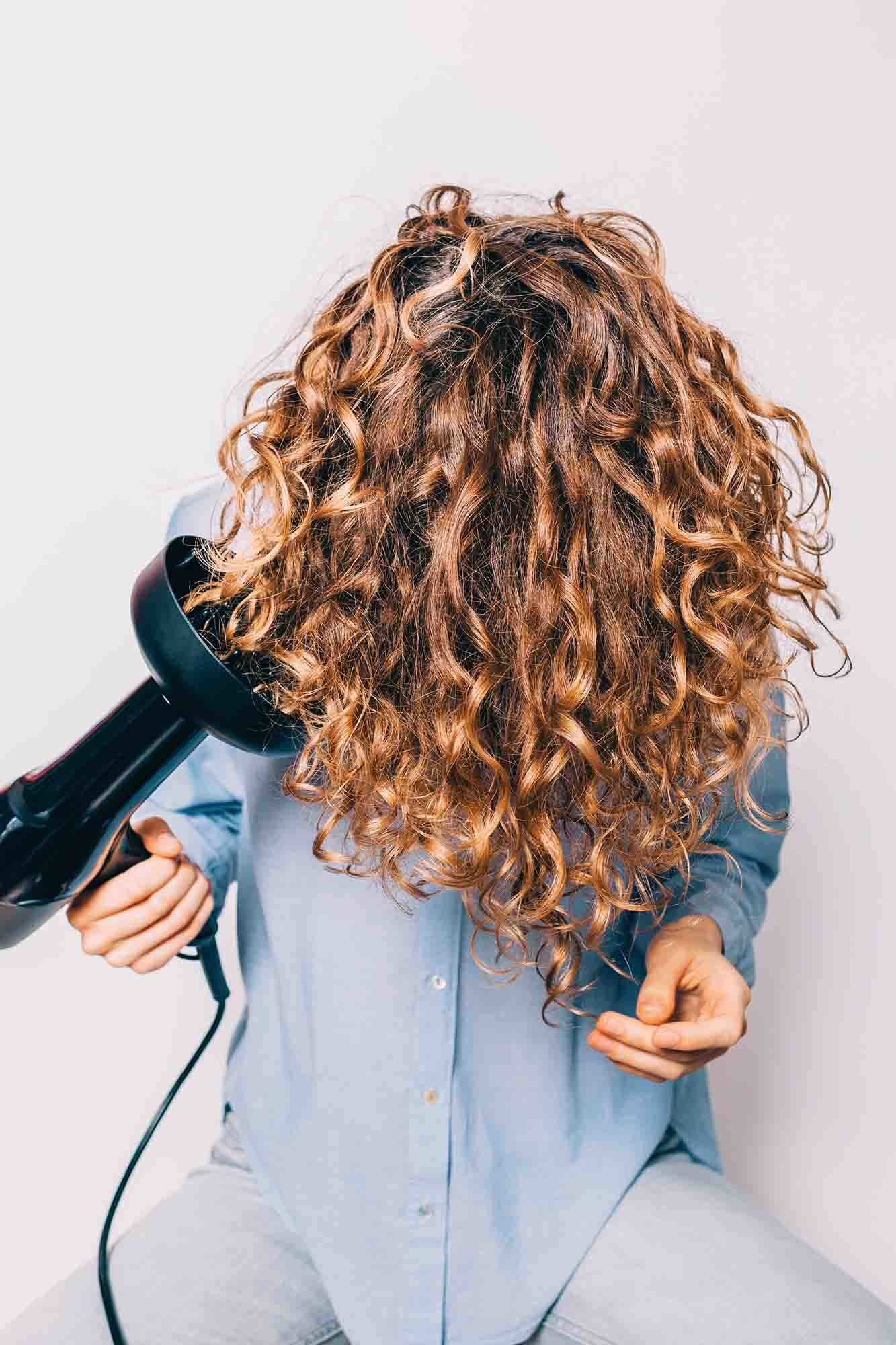 wanita dengan rambut ikal berwarna cokelat sedang mengeringkan rambut dengan diffuser hairdryer