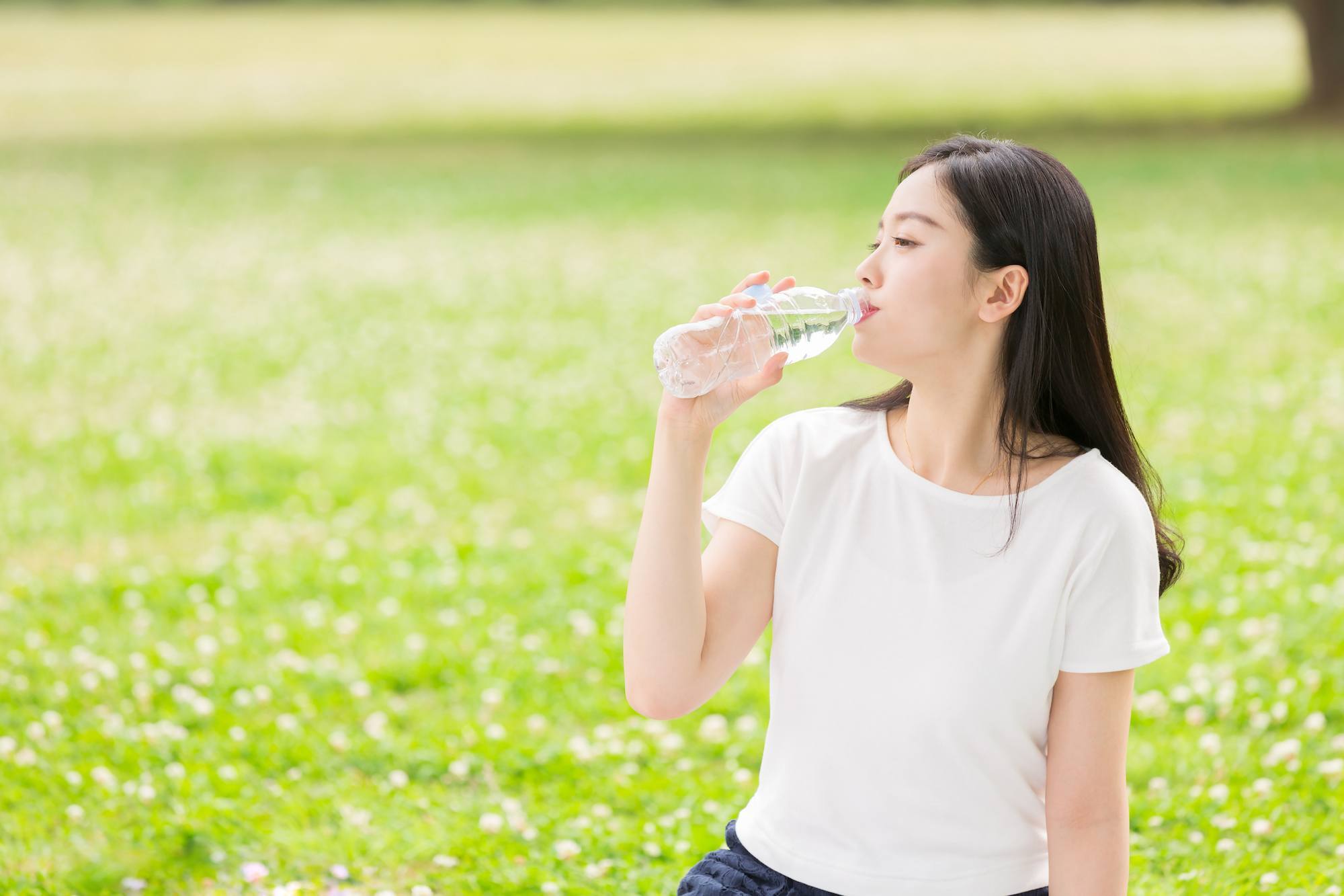 Wanita asia dengan rambut hitam panjang minum sebotol air putih di atas lapangan hijau.