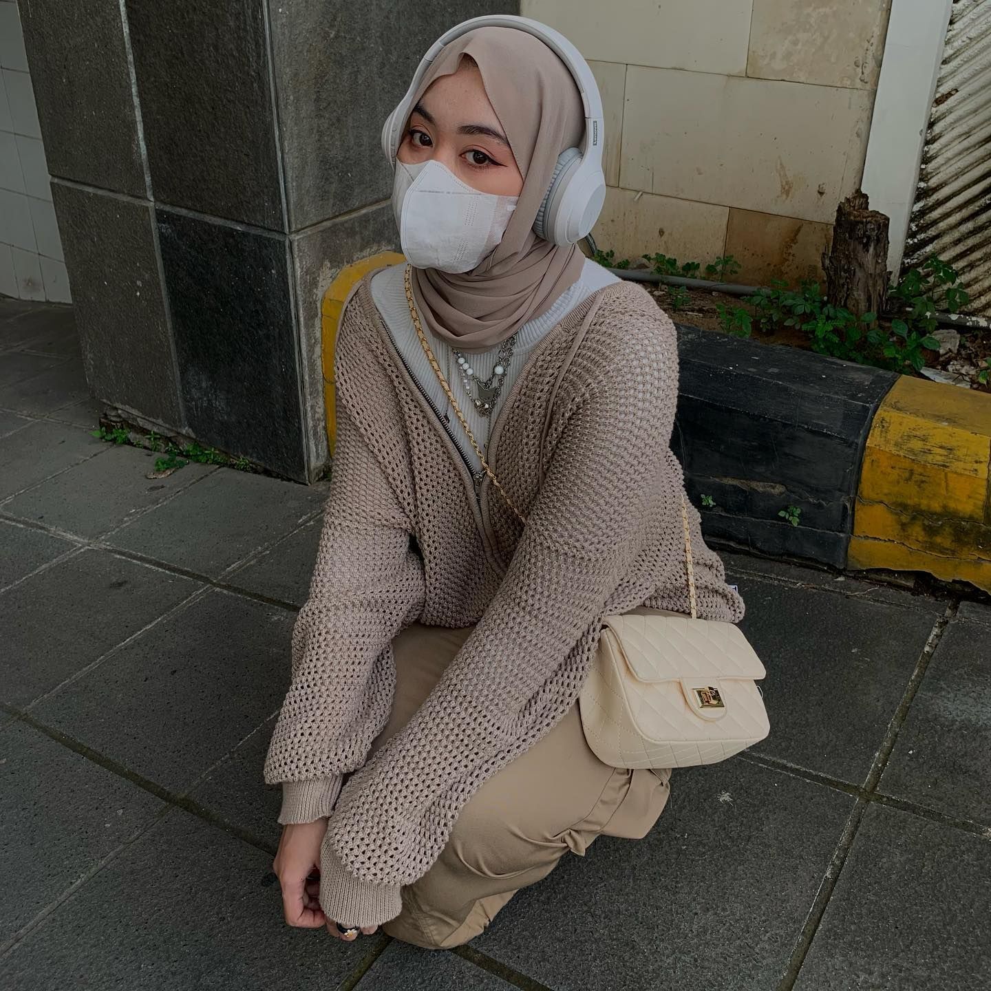 Wanita berhijab sedang duduk jongkok mengenakan outfit dan hijab pashmina warna cream dan headphone warna putih.