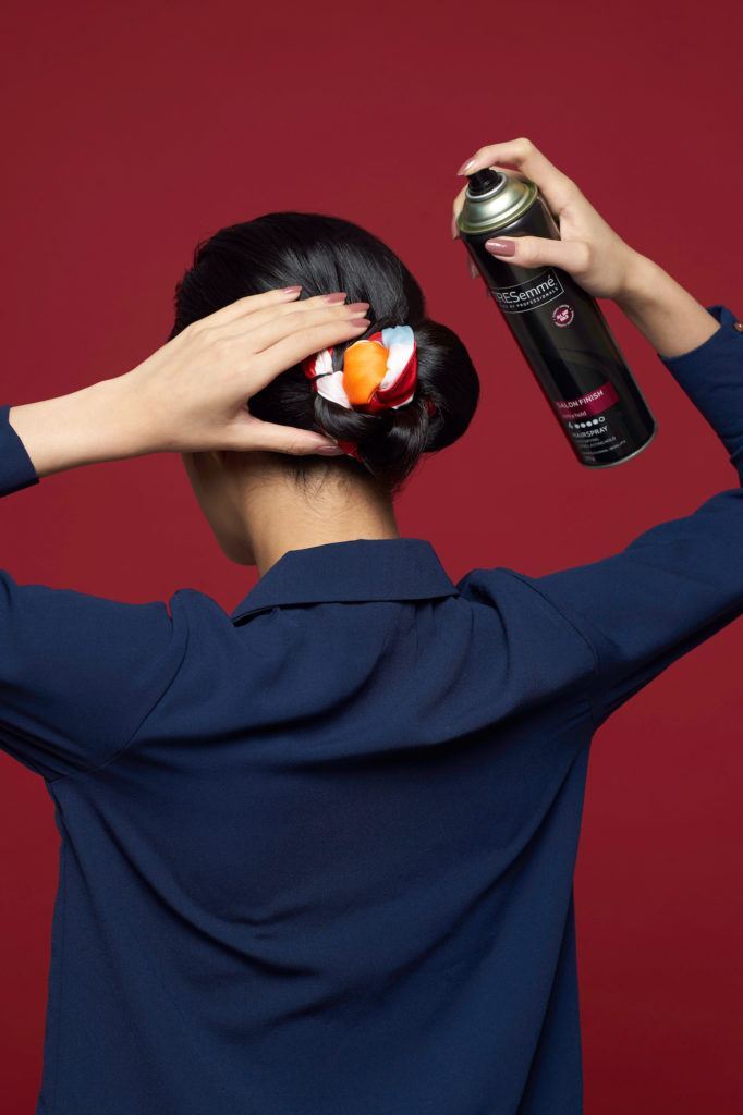 Wanita asia dengan rambut hitam menyemprotkan hairspray pada rambutnya – cara menata scarf bun
