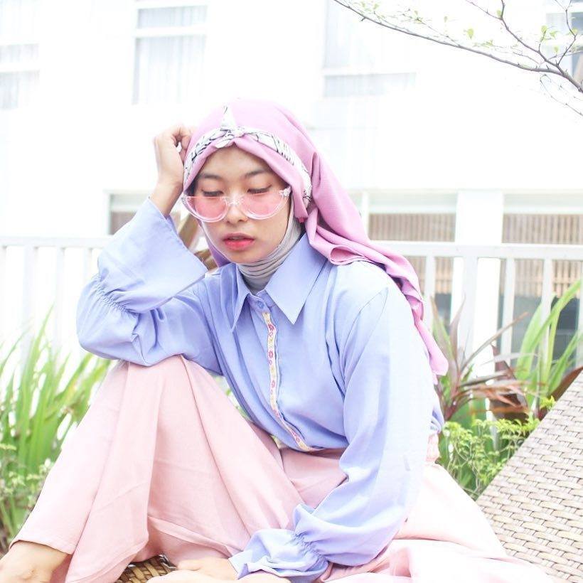 Wanita berhijab unik dan kaca mata pink menopang kepala dengan tangan di depan rumah.