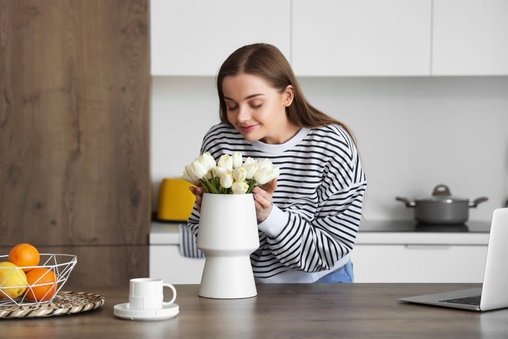 หญิงสาว ผมยาว ก้มดม ดอกไม้สีขาว ในแจกัน มีภาชนะ ใส่ ผลส้ม วางบนโต๊ะ