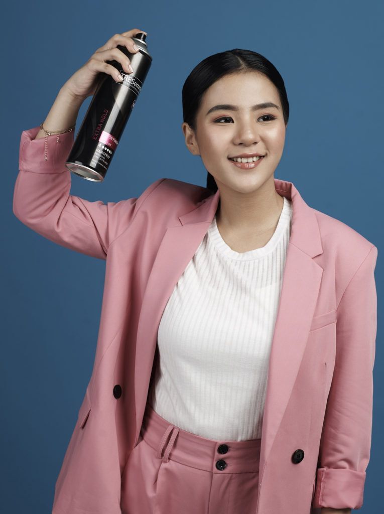 ผู้หญิงผมยาวสีดำ มัดผมหางม้า สวมสูทสีชมพู กำลังฉีดสเปรย์แต่งผม