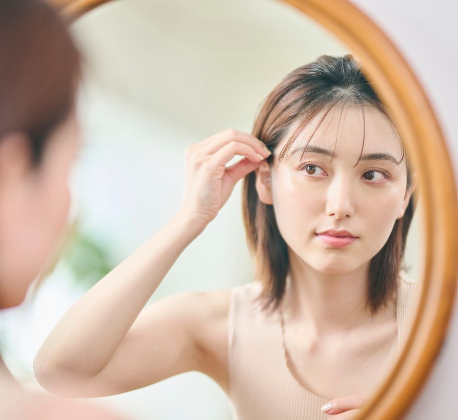 Woman with short hair and thin bangs looking at a mirror.