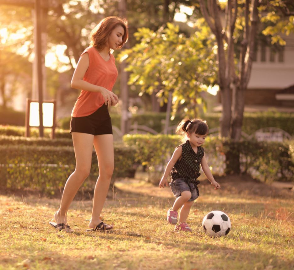 Asian mom and daughter playing soccer outdoors.