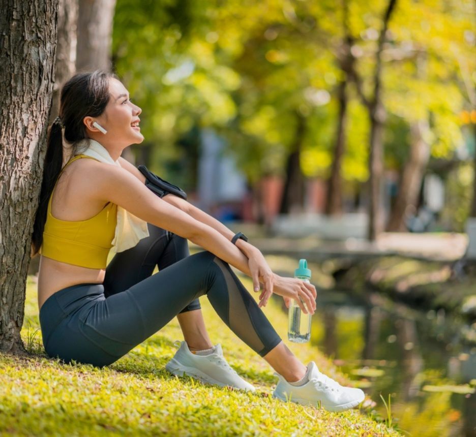 Young Asian female in fitness clothes relaxes by a park’s stream after a workout.