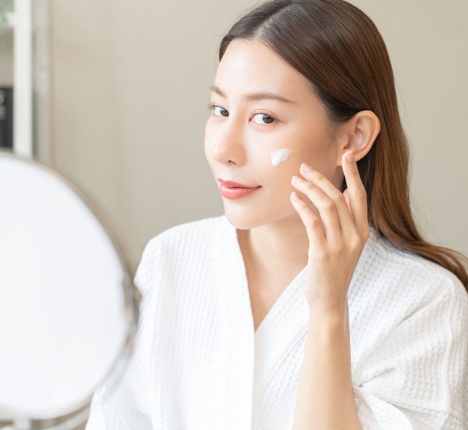 cách chăm sóc da thường thoa kem chống nắng cô gái khoác áo choàng trắng ngồi thoa kem trước gương tròn