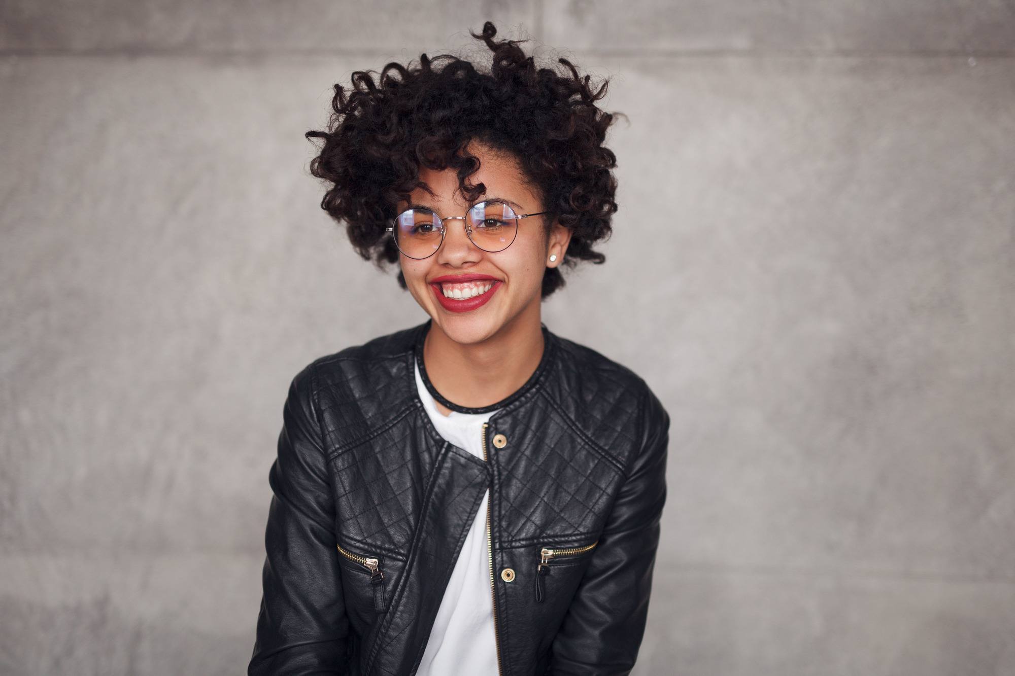 self-isolation hairstyles: woman in glasses and leather coat smiling and looking away on concrete wall background