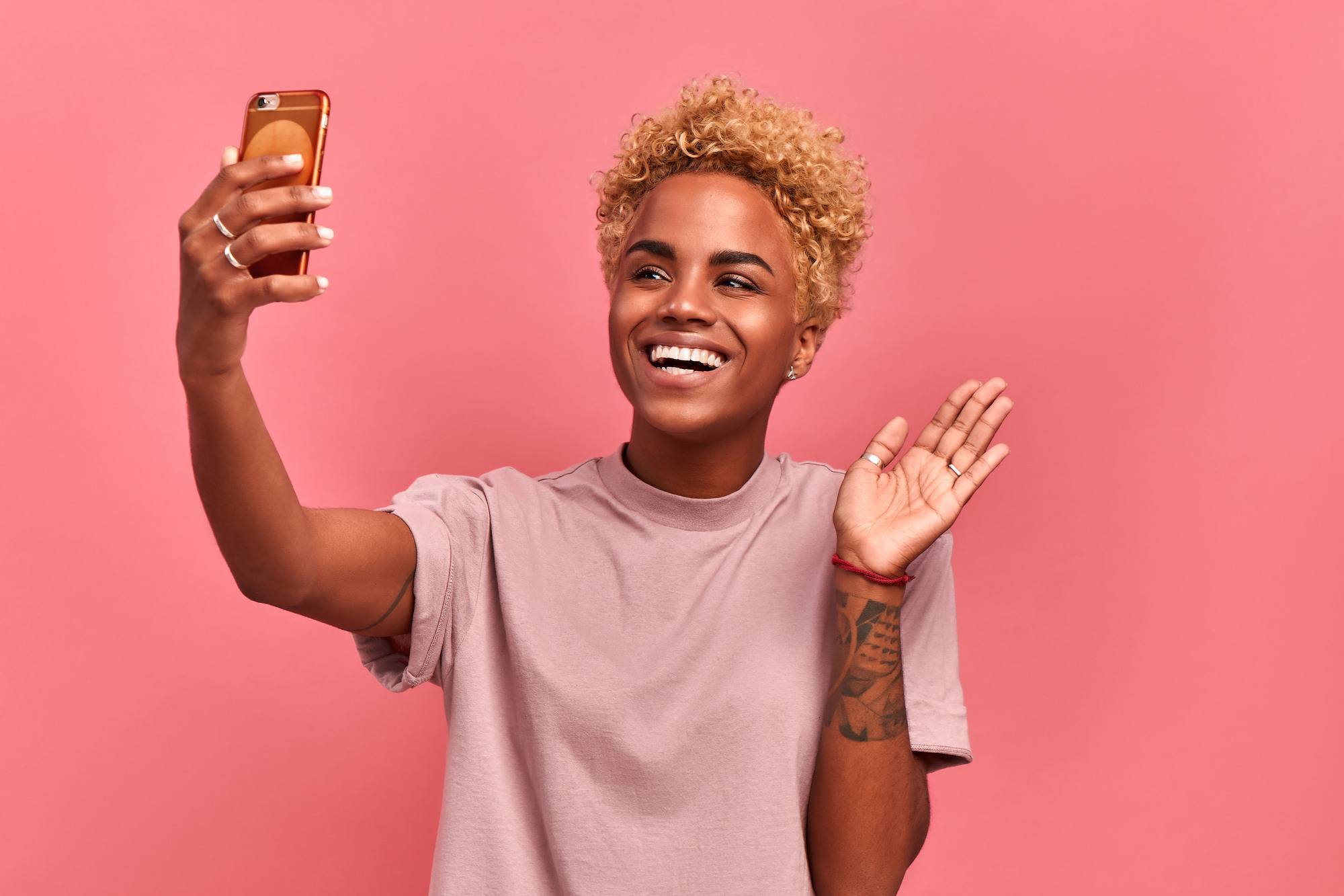 Glad dark skinned happy woman with twa blonde Afro hairstyle, holds modern mobile phone in front of face, waves palm in camera, makes video call, dressed in casual wear, isolated over pink background.