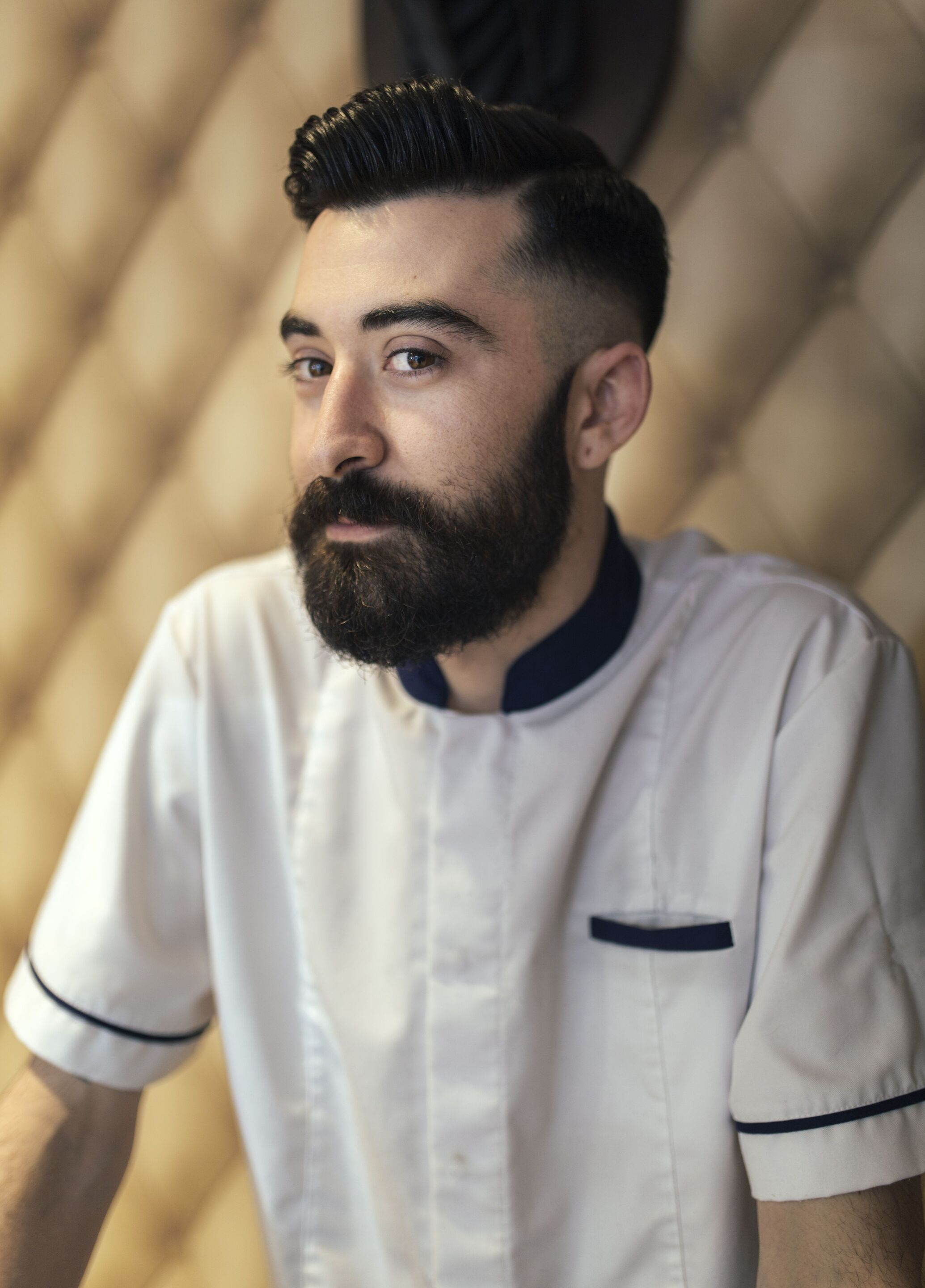 Man with dark brown hair styled in a pompadour with an undercut. Man also has a full beard and moustache and he is wearing a barber top.