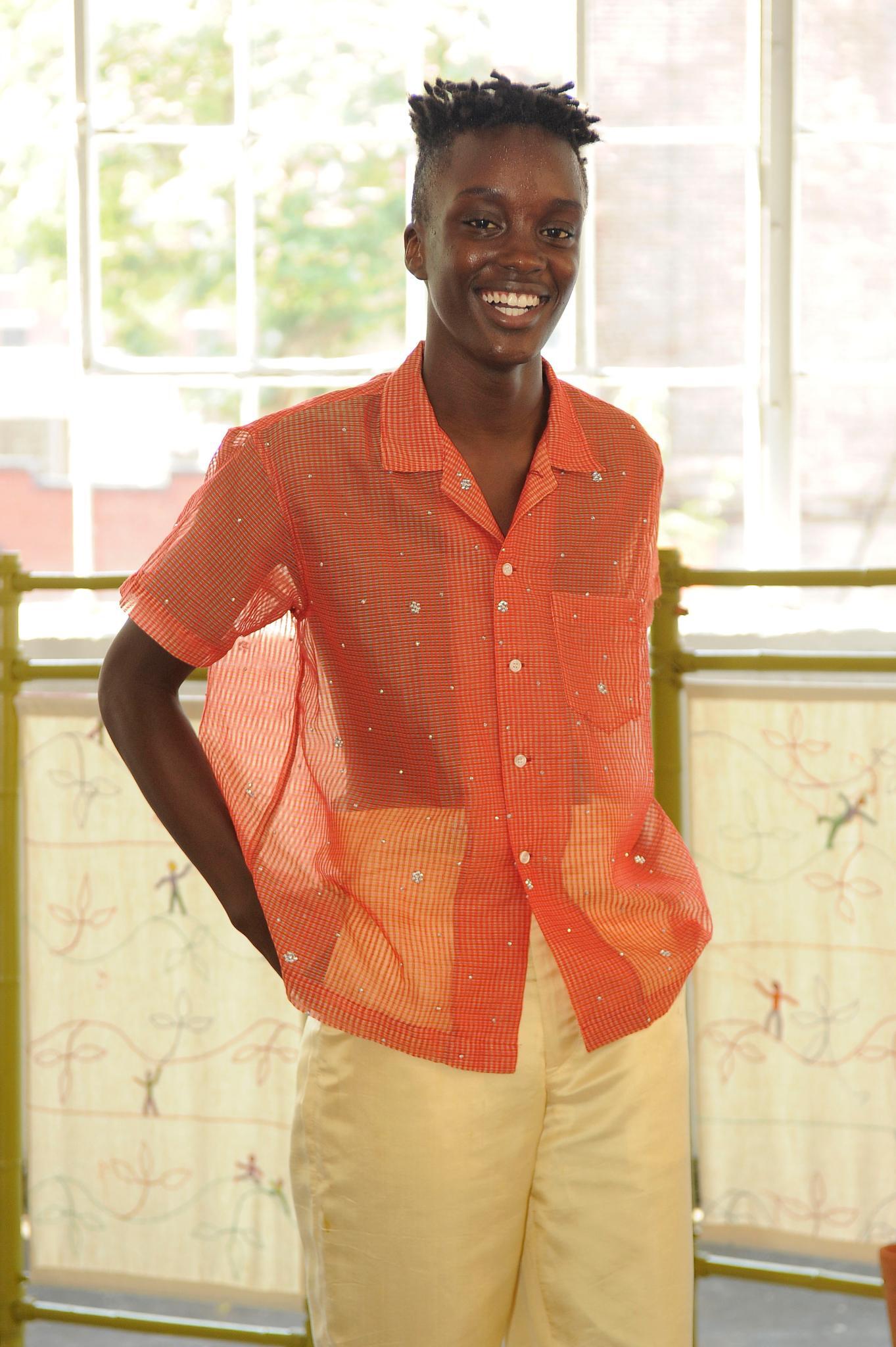 Male model at Bode SS19 show with sponge twists and undercut hair wearing a orange sheer shirt and trousers smiling at the camera.