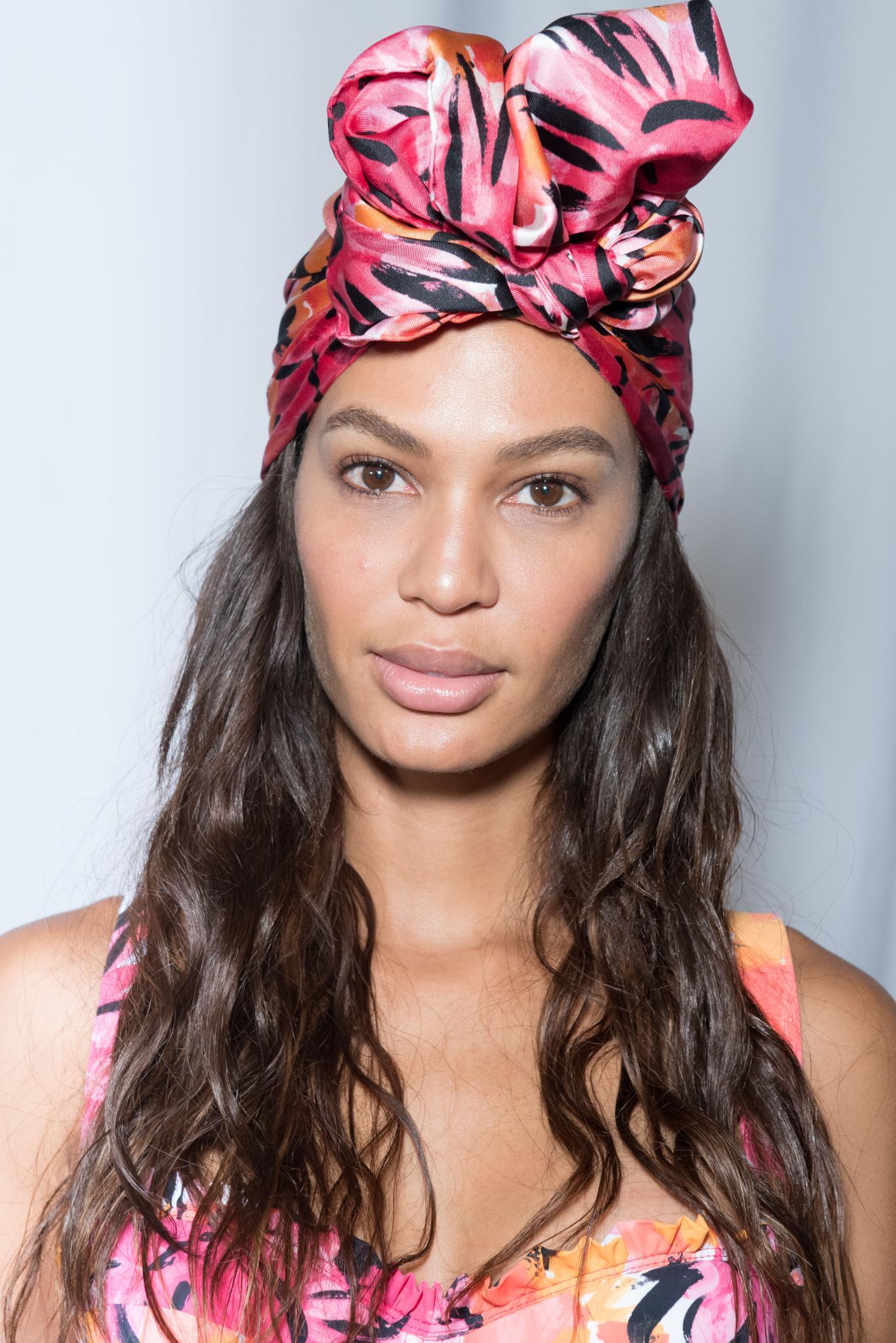 New hairstyle: Joan Smalls at Michael Kors SS19 show with long wavy brown hair wrapped in a pink headscarf.