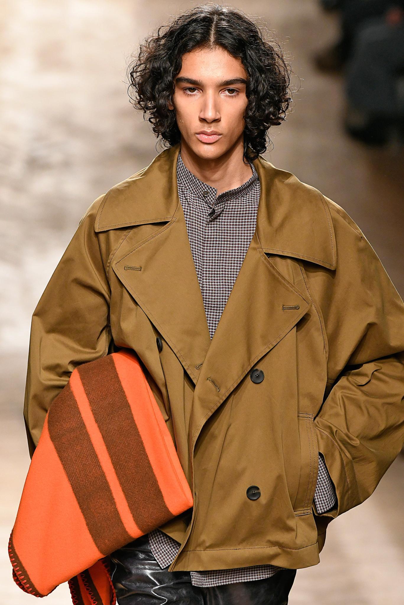 Man on runway with shoulder length curly brown hair wearing a dark camel jacket.