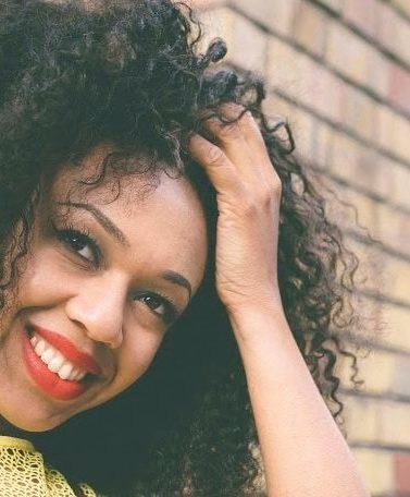African hair: Woman touching her naturally curly dark brown shoulder length hair while leaning against a brick wall. Woman is wearing a yellow top and bold coral lipstick.
