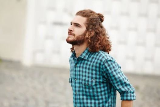 Man with curly auburn shoulder length hair in a half-up top knot wearing a green and white check shirt.
