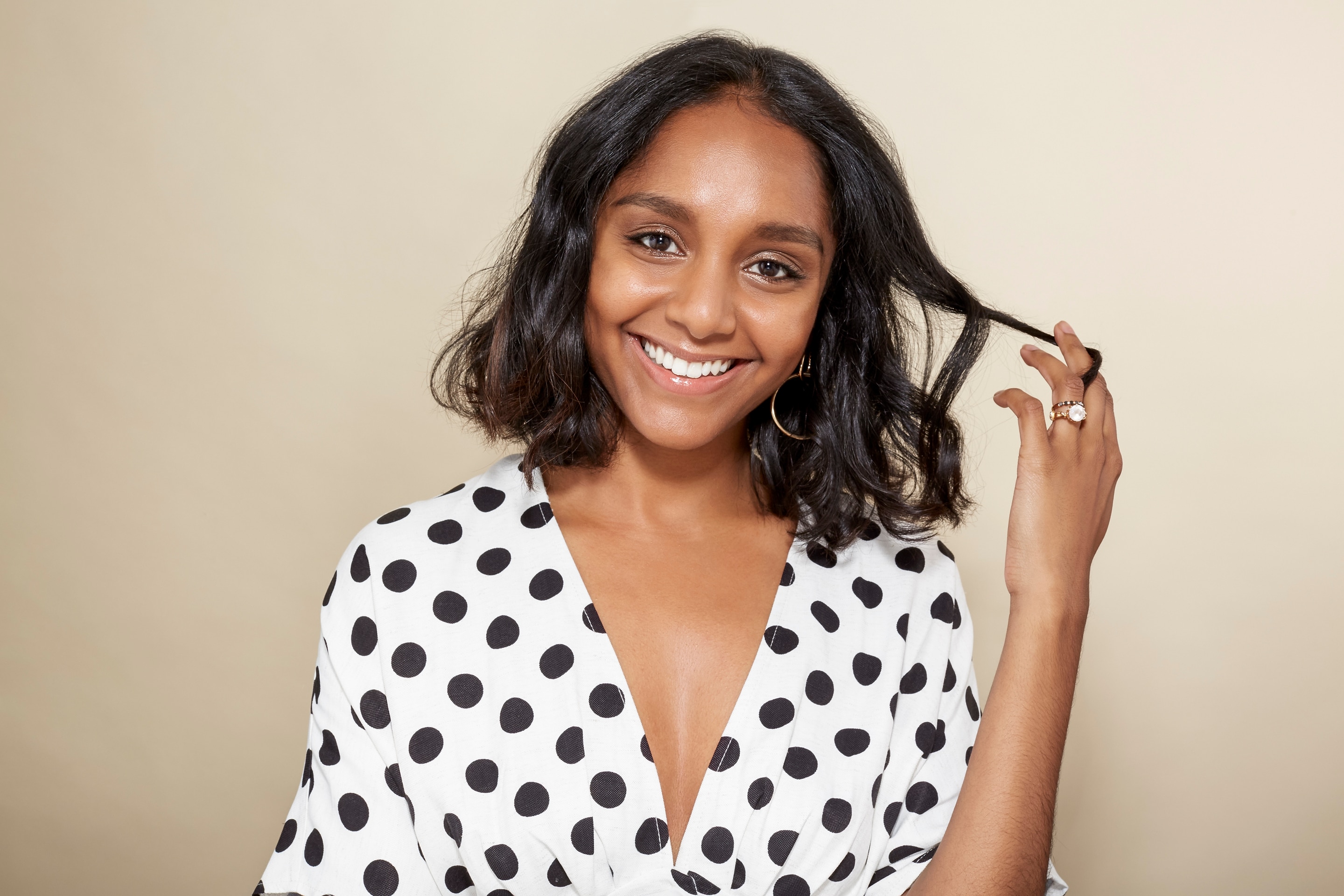 Short hairstyles for thick hair: Woman with a wavy long bob touching her hair. Woman is wearing a white top with black polka dots and smiling at the camera.