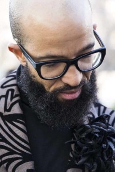 Man with shaved bald head and long dark brown beard wearing glasses and black strip blazer in street style image.