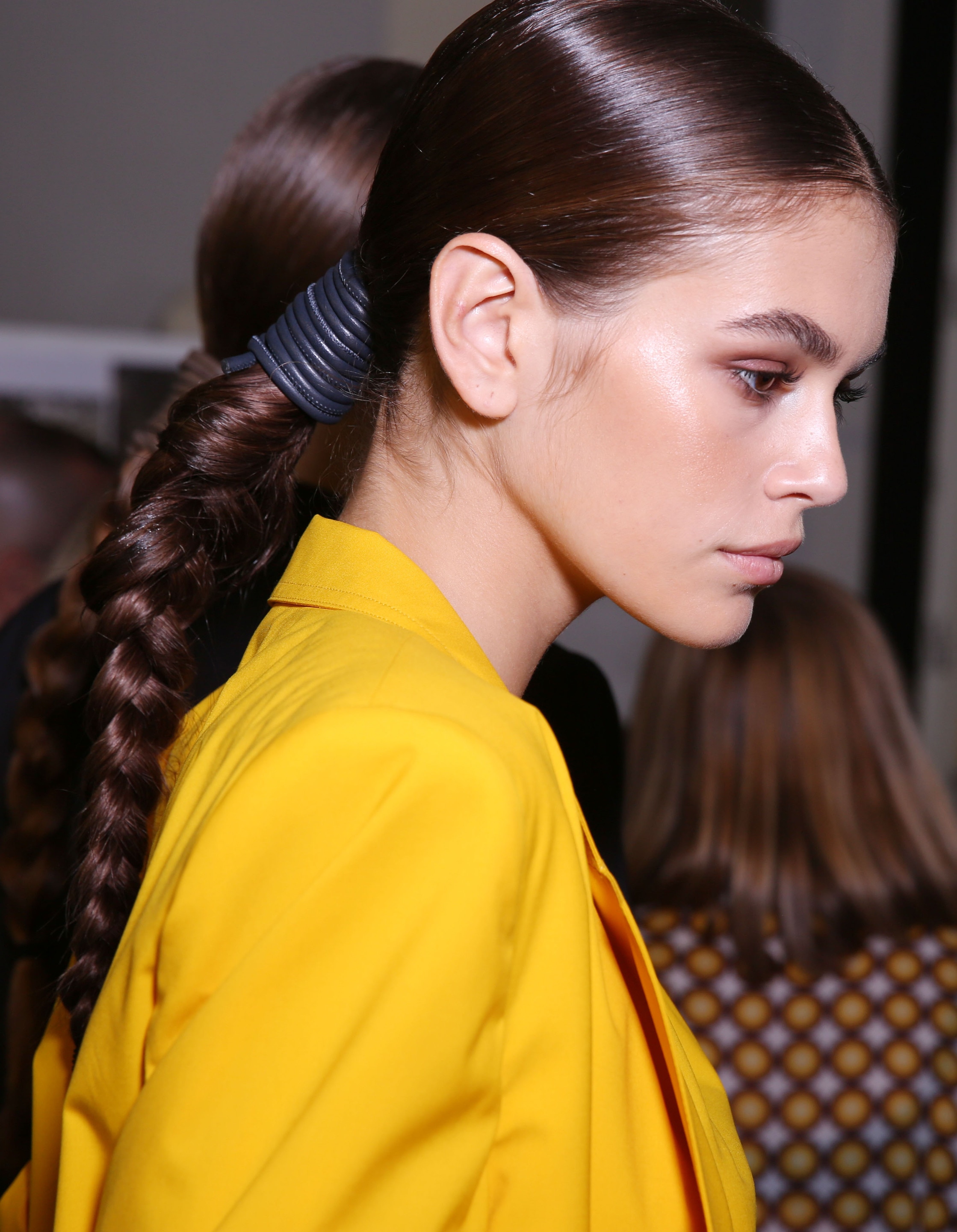 Kaia Gerber with brown hair in low ponytail braid in backstage runway image wearing a yellow blazer.