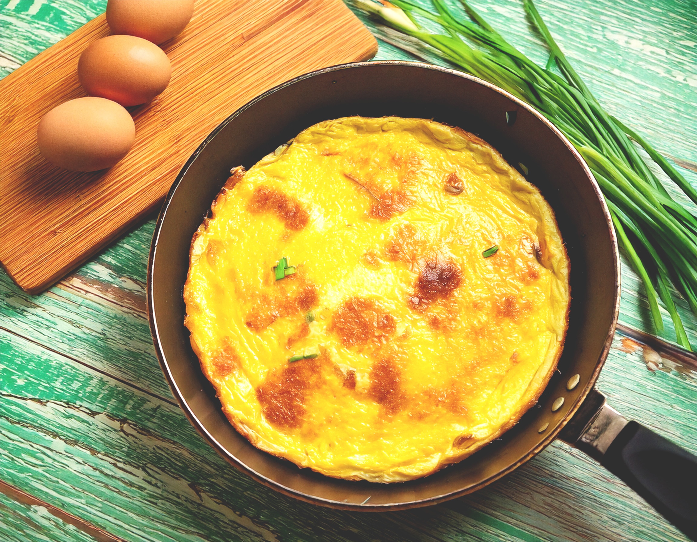 Einfaches Omelette in einer Pfanne, Eier und Schnittlauch liegen daneben auf einem Brett