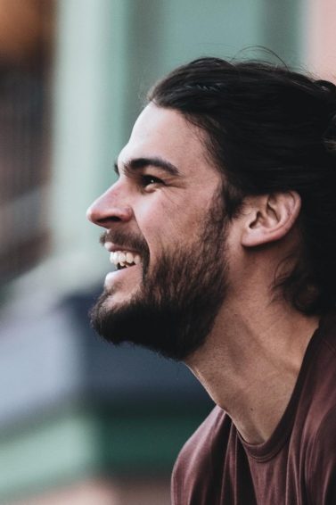 Homem de cabelo longo preso e camiseta marrom sorri ao ar livre