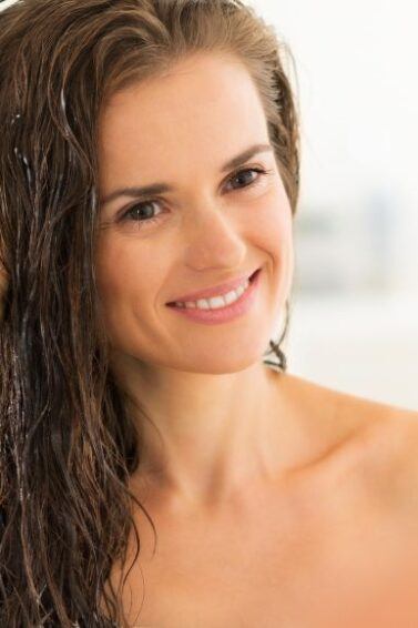 Mulher com as mãos no cabelo castanho sorrindo ao se olhar no espelho