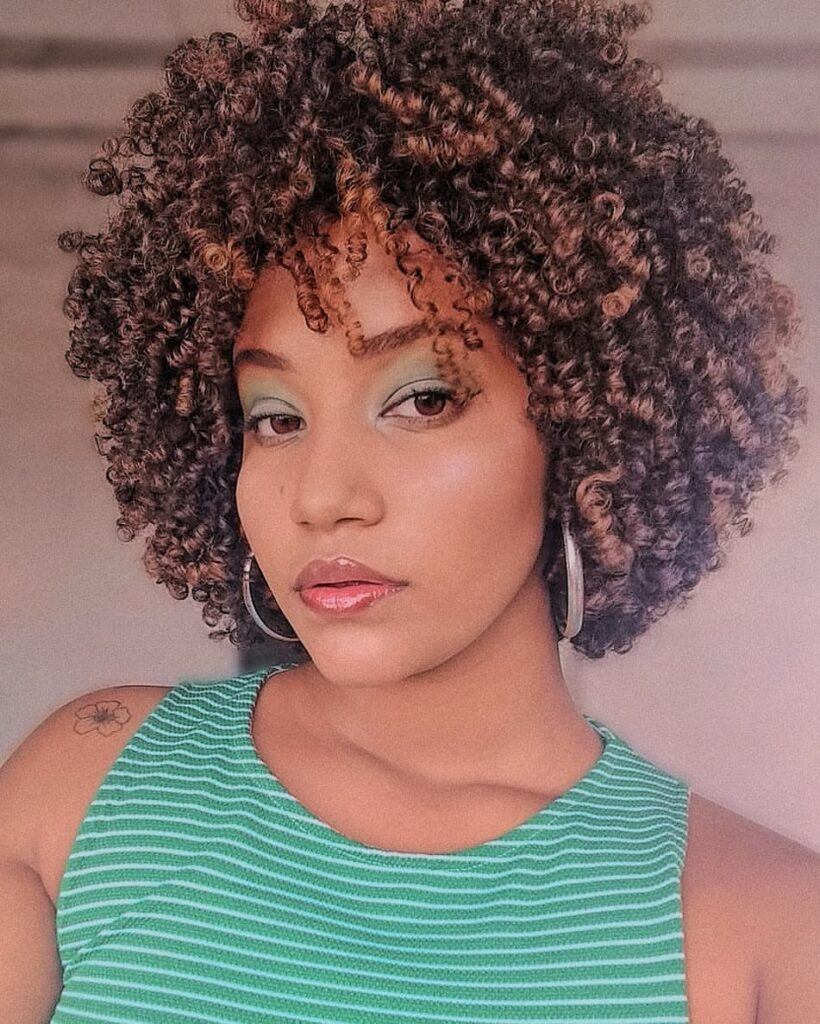 Mulher posa com top verde listrado, brinco argola e cabelo cacheado curto 3c castanho claro com luzes