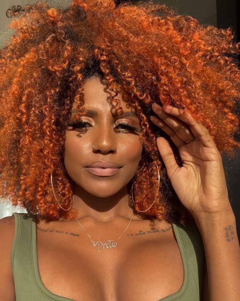 Mulher ruiva posa de cabelo cacheado 3c com franja e top verde, com a mão no rosto