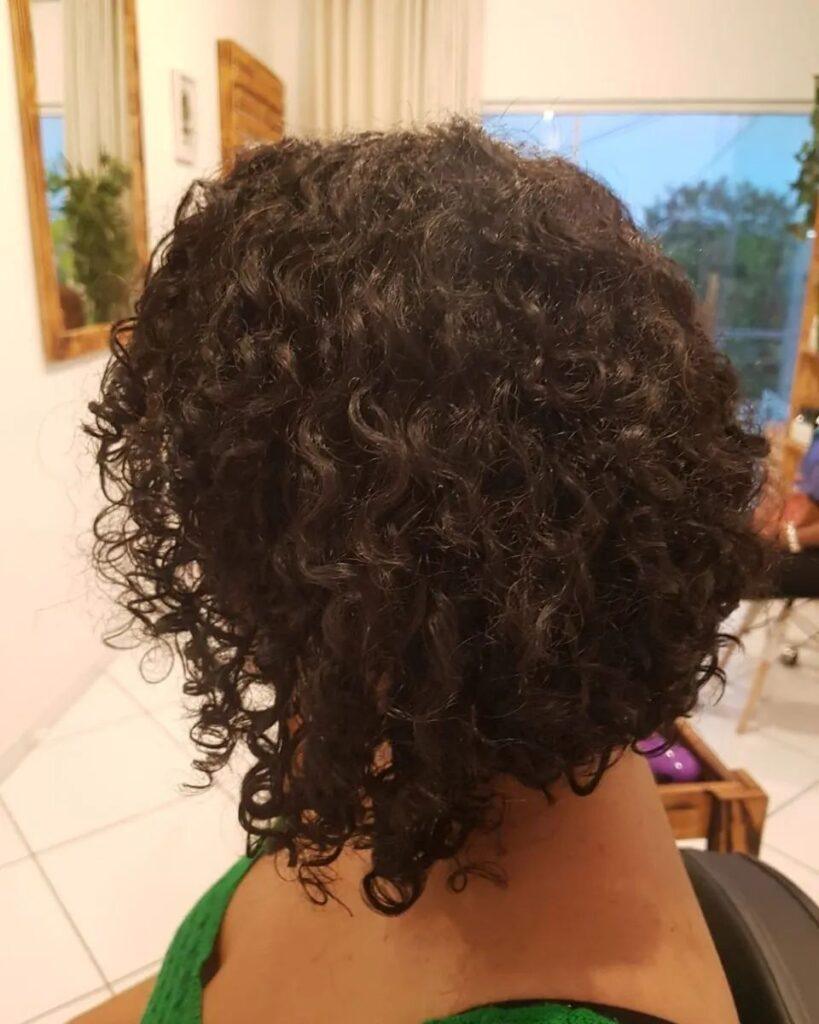 Mulher posa de perfil no salão, destacando o corte de cabelo cacheado 3c curto assimétrico estilo chanel de bico
