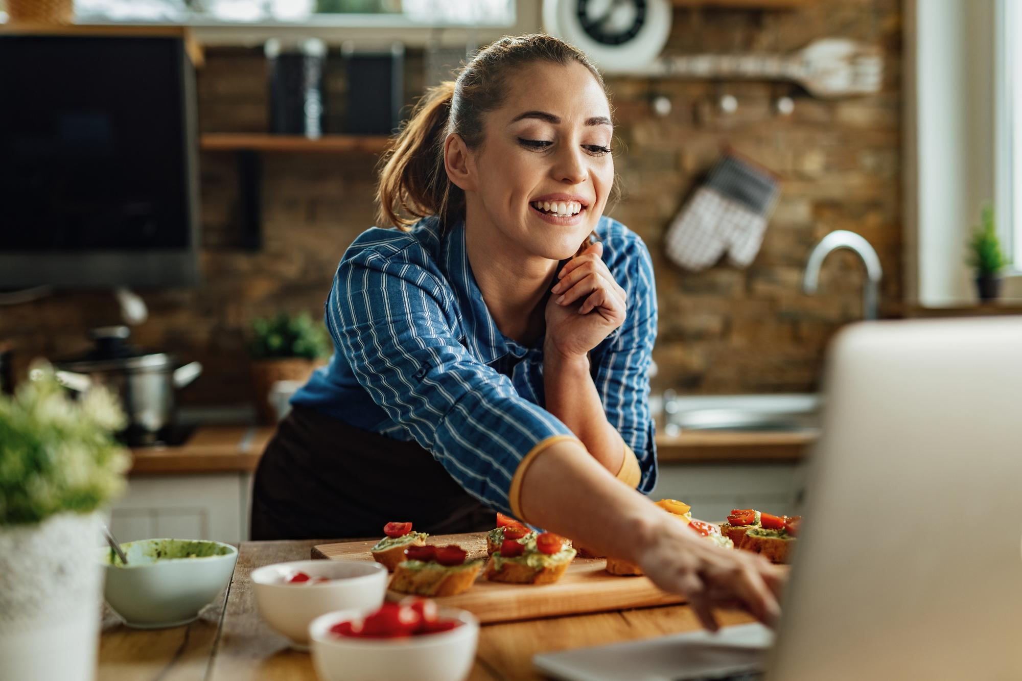 Mulher consulta seu laptop enquanto prepara receita na cozinha durante férias em casa