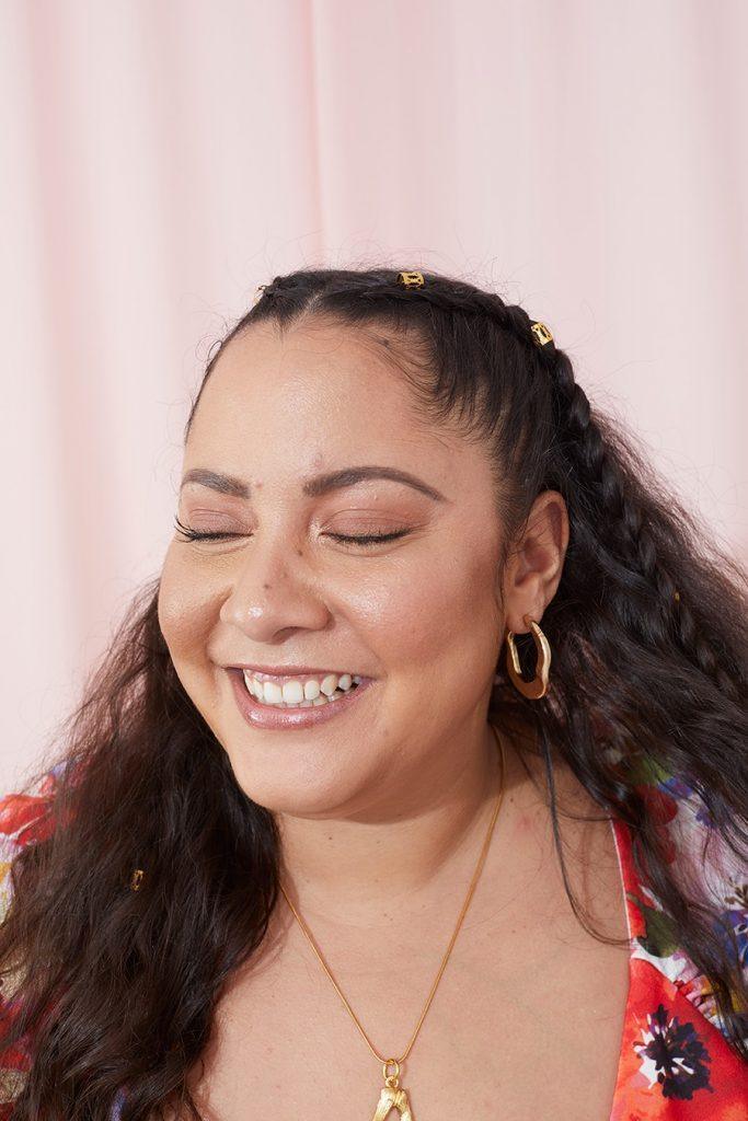Mulher com blusa florida e cabelo semipreso com trancinhas sorri de olhos fechados