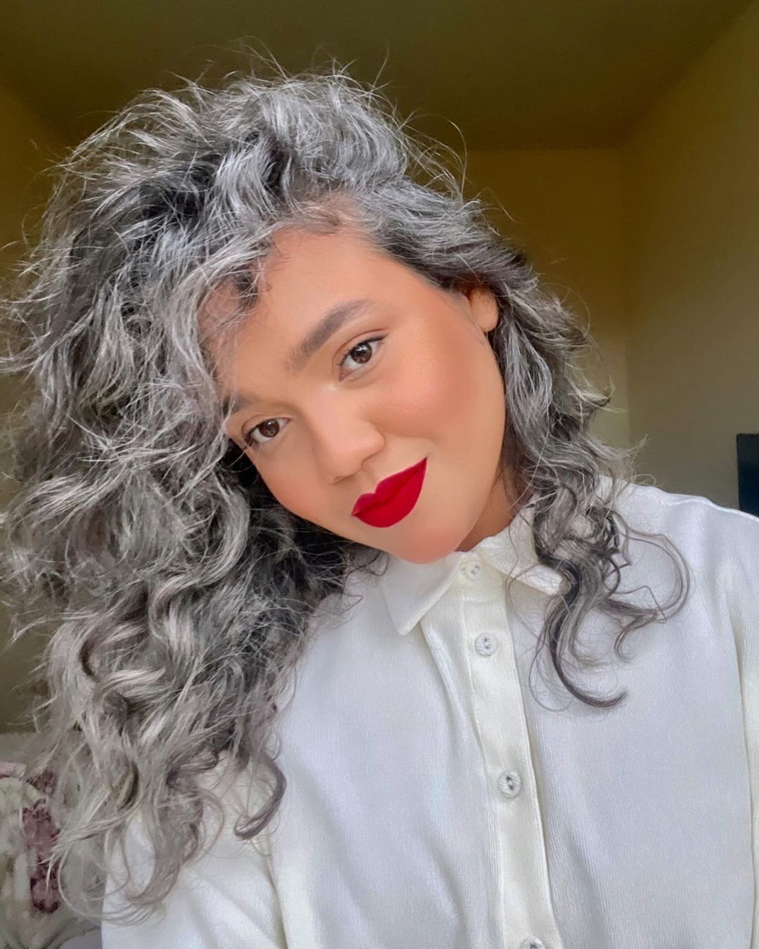 mulher jovem de cabelo grisalho posa de camisa branca e batom vermelho, evidenciando o cabelo grisalho cacheado
