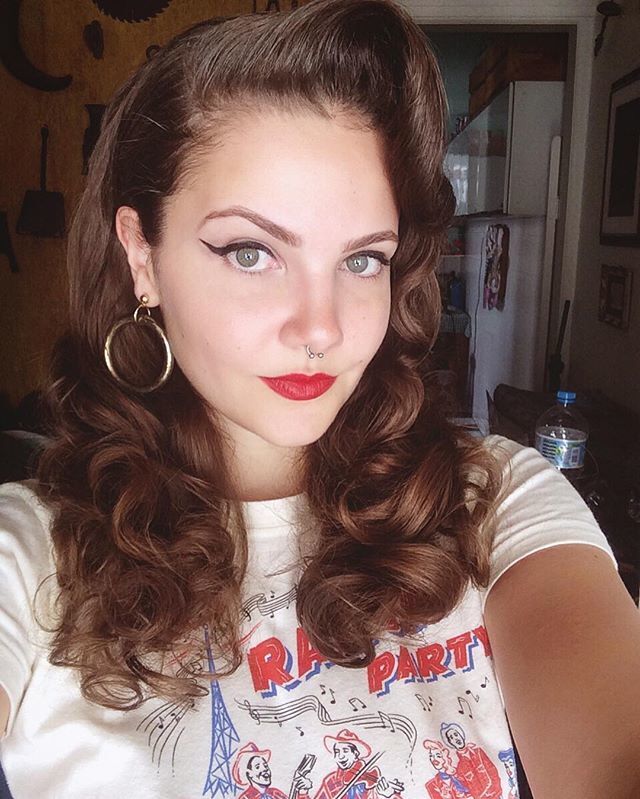 Mulher de cabelos cacheados castanhos e topete intage faz selfie. Ela usa brinco de argola, batom vermelho e delineador gatinho