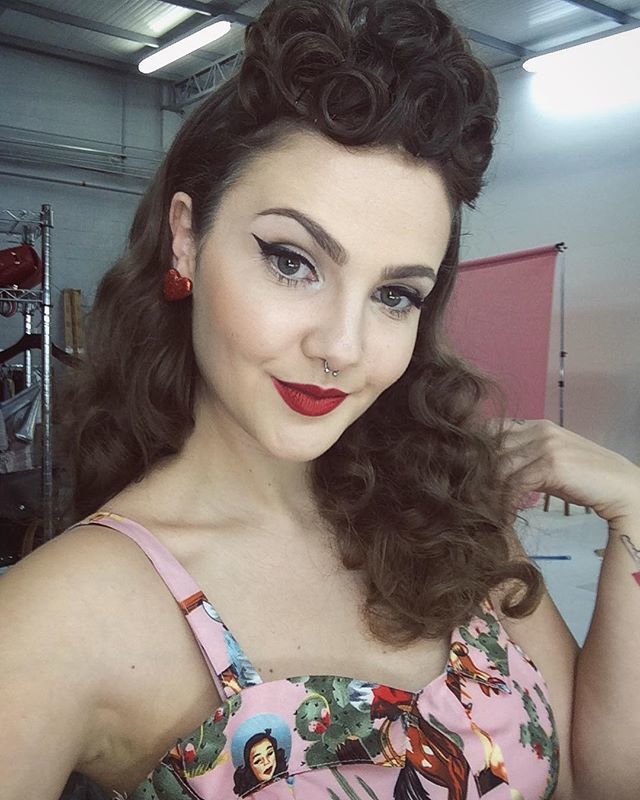Mulher de cabelos cacheados castanhos e topete vintage faz selfie. Ela usa brinco de coração, batom vermelho e delineador gatinho