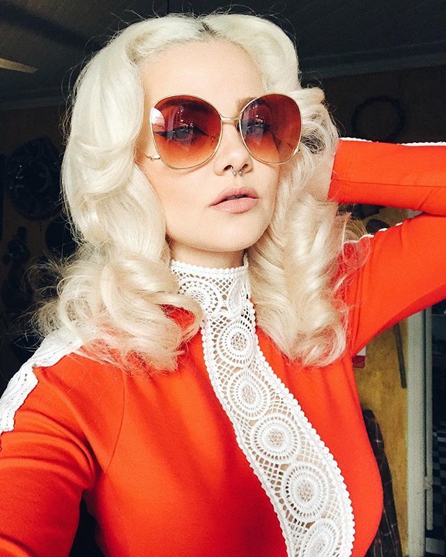 Mulher de cabelo platinado e com cachos, óculos escuros grandes, batom claro posa para selfie com a mão na cabeça