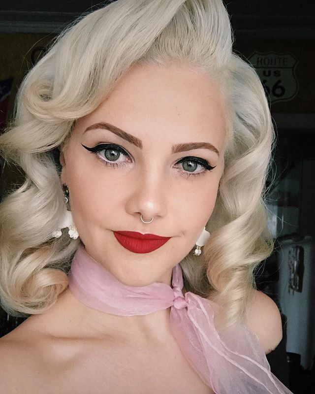 mulher com cabelo loiro platinado e penteado vintage posa para a câmera. Ela usa lenço rosa amarrado no pescoço, batom vermelho e delineador gatinho