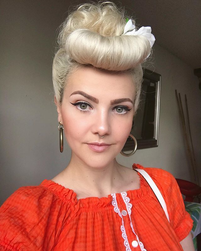 mulher com cabelo loiro platinado e penteado vintage posa para a câmera. Ela usa roupa laranja,, brinco de argola e delineador gatinho