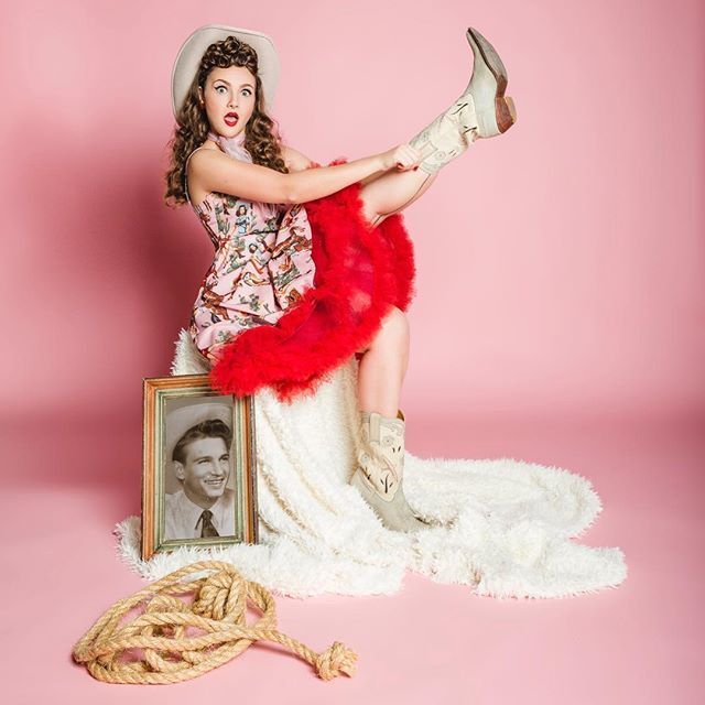 modelo posa segurando a bota de cowboy com a perna para cima, sentada em um caixote coberto por uma manta. Ela usa vestido rosa, com babados vermelhos, chapéu de cowboy. Na cena existe um quadro com foto preto e branco de um homem e uma corda