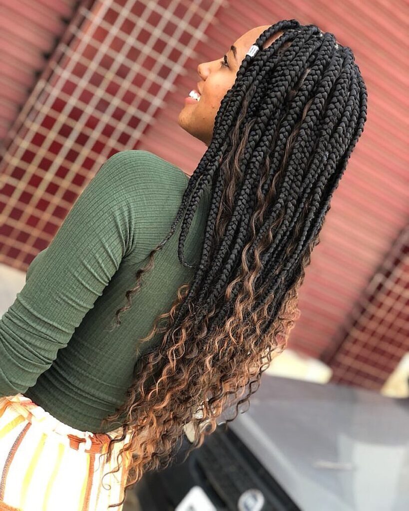 Mulher com blusa verde posa de lado, destacando o cabelo longo com tranças goddess braids no cabelo castanho com luzes nas pontas