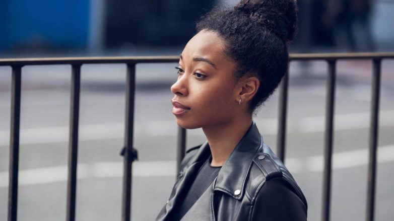 Mulher negra usa cabelo preso num coque, jaqueta de couro e blusa preta. Ela ilustra matéria sobre como deixar o cabelo crespo mais solto