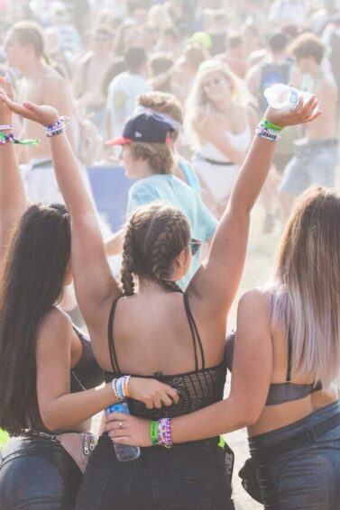 Imagem de 3 amigas se abraçando em festival de música com outras pessoas em volta no ambiente ao ar livre cheio