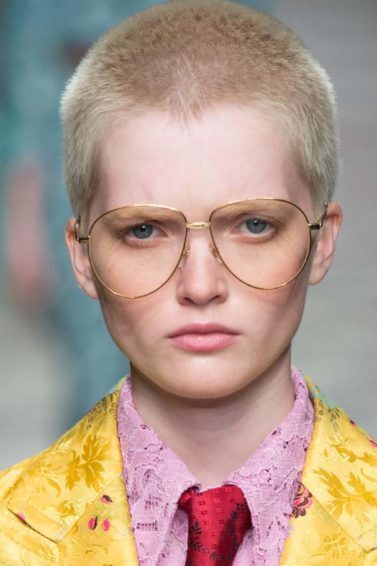 Mulher usa terno amarelo, camisa rosa, gravata vermelha, óculos tipo aviador e buzz cut platinado