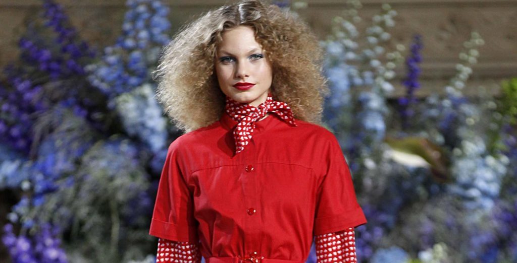 Mulher loira usa cabelo volumoso indefinido e veste roupa vermelha, na passarela